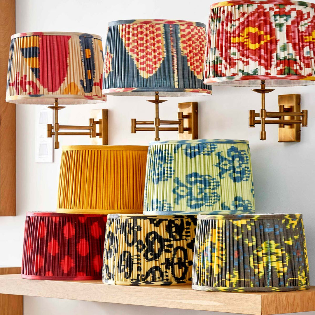 Collection of Mekhann's hand-pleated silk ikat lampshades in various vibrant colours and floral patterns displayed on wooden shelves.