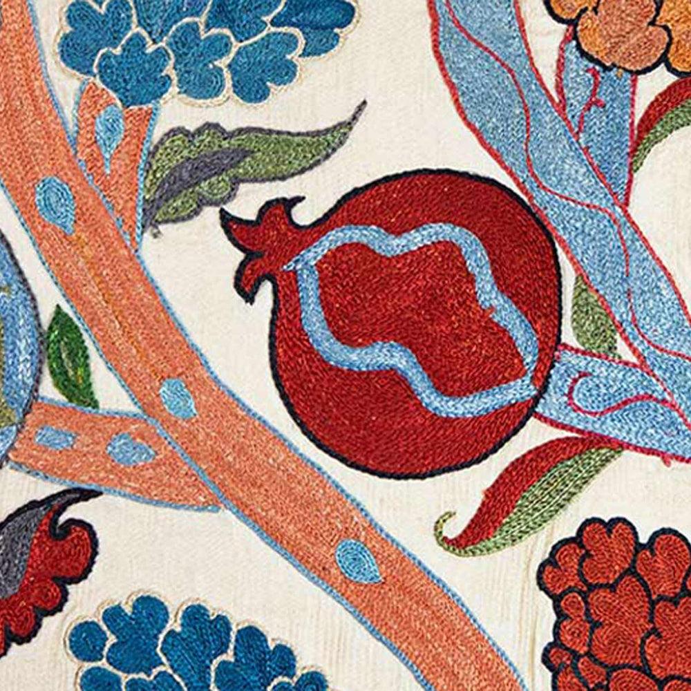 Close up view of Mekhann's cream Grapes and Pomegranates embroidered cushion, where the focal point is a red hand embroidered pomegranate with strands of blue pattens growing though it.