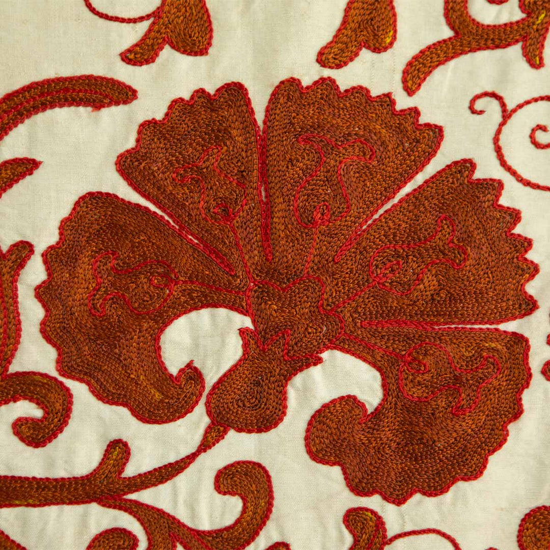 Close up view of Mekhann's cream carnations throw, showing in detail the texture of the hand embroidered, orange carnations motif, with red details running through, all set on a background of cream coloured silk. 