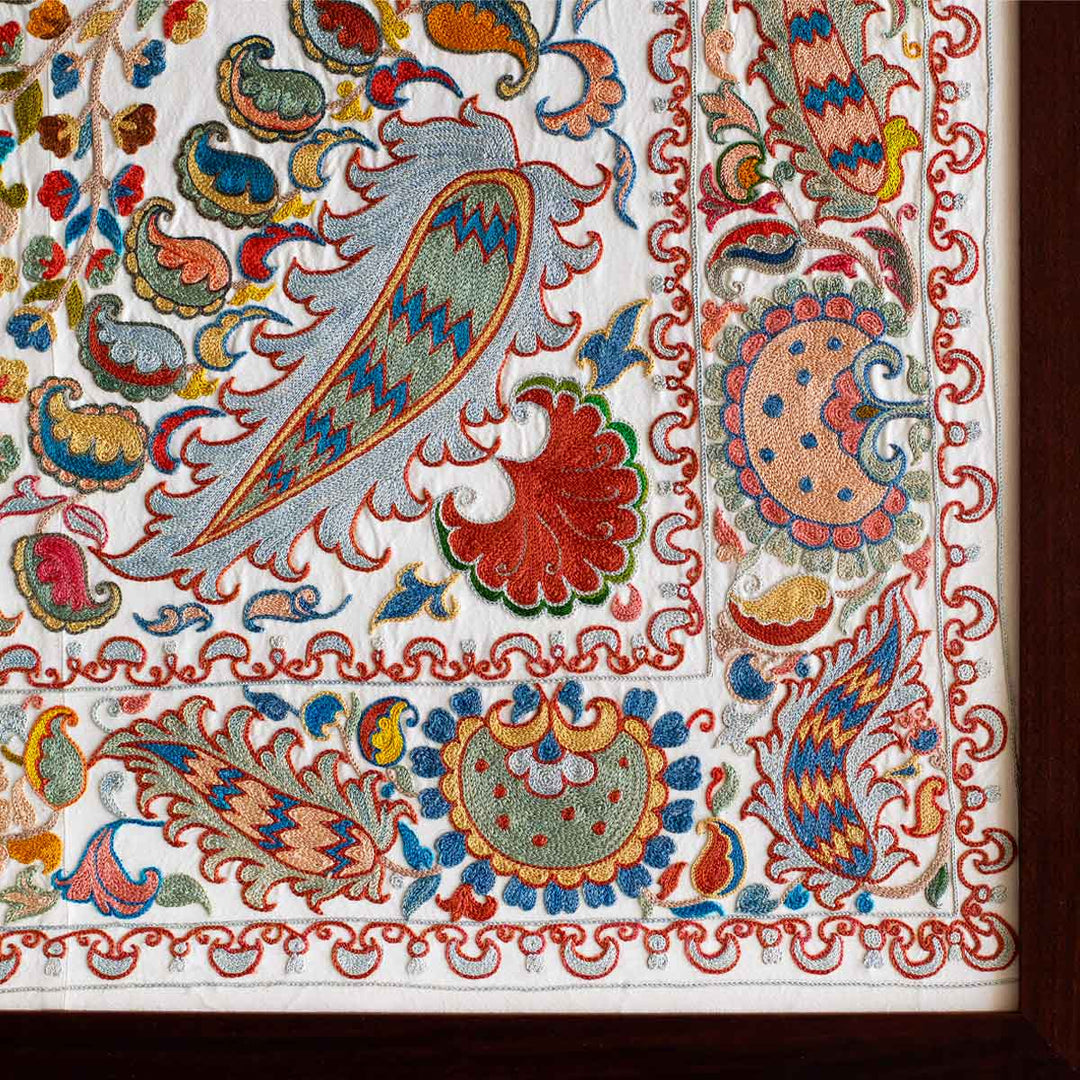 Corner view of cream silk, multicoloured embroidered framed artwork, showing where the silk art work ends and the dark brown frame begins.