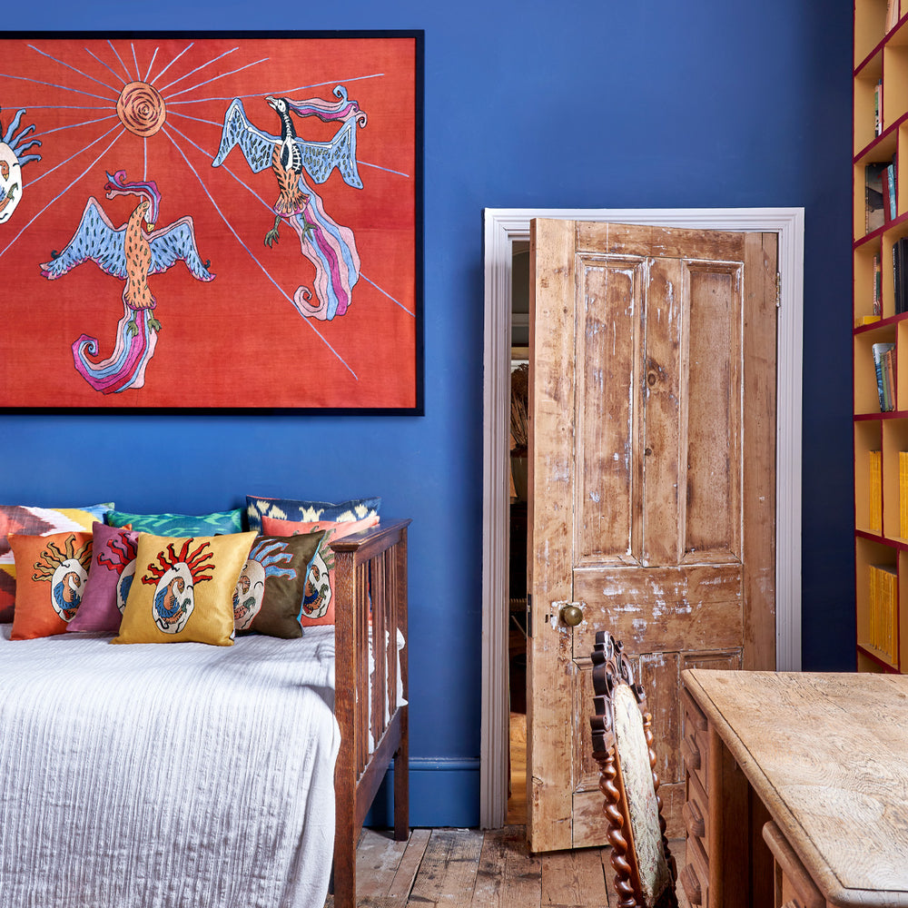 In use view of Mekhann's maroon silk hand embroidered phoenix artwork, showing the silk artwork on a blue wall, displaying how the piece might interact with someones interiors.