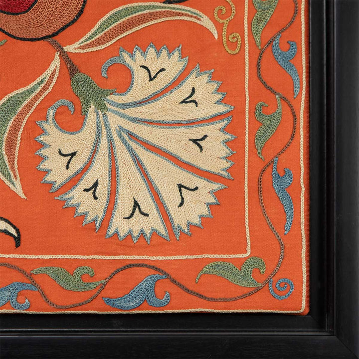 Corner view of Mekhann's orange silk floral artwork, showing a cream hand embroidered carnation in the bottom corner set against the orange silk. We can also see the black frame up close.
