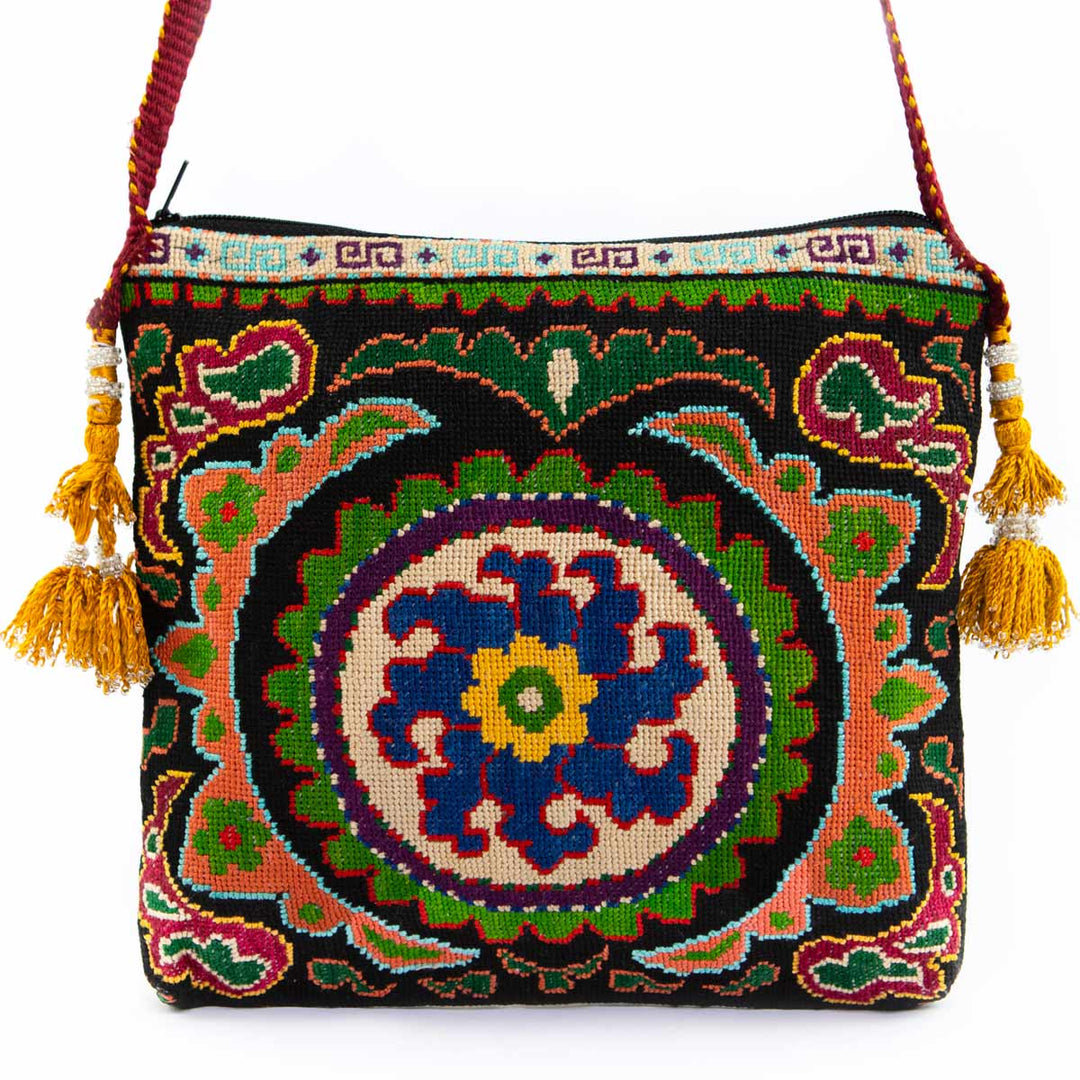 Front view of Mekhann's black and multicoloured floral embroidered cross-body bag, displaying it's yellow side tassels and red strap.