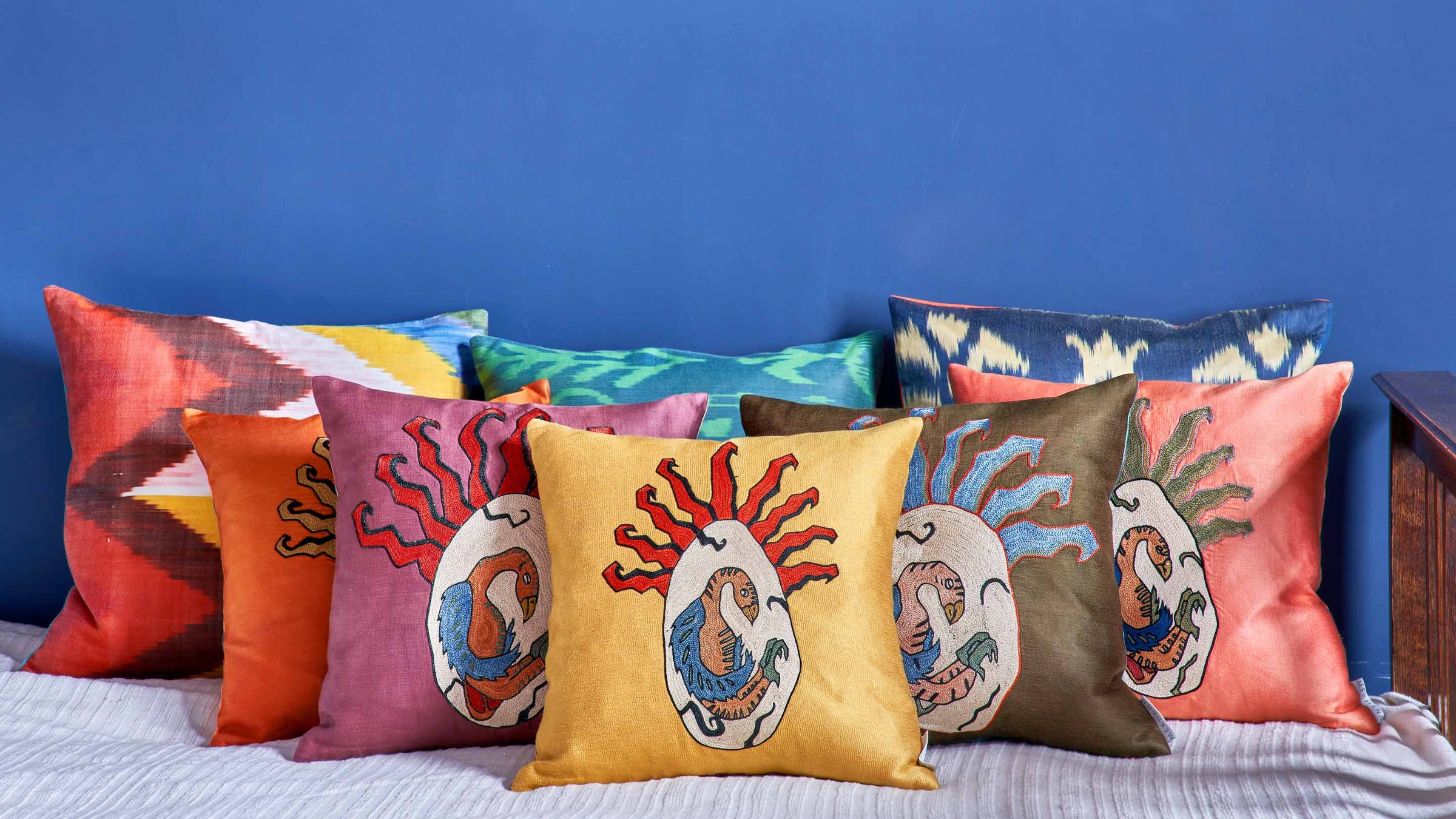 Mekhann Phoenix Egg embroidered cushions arranged on a bed with ikat cushions, styled against a bold blue wall.