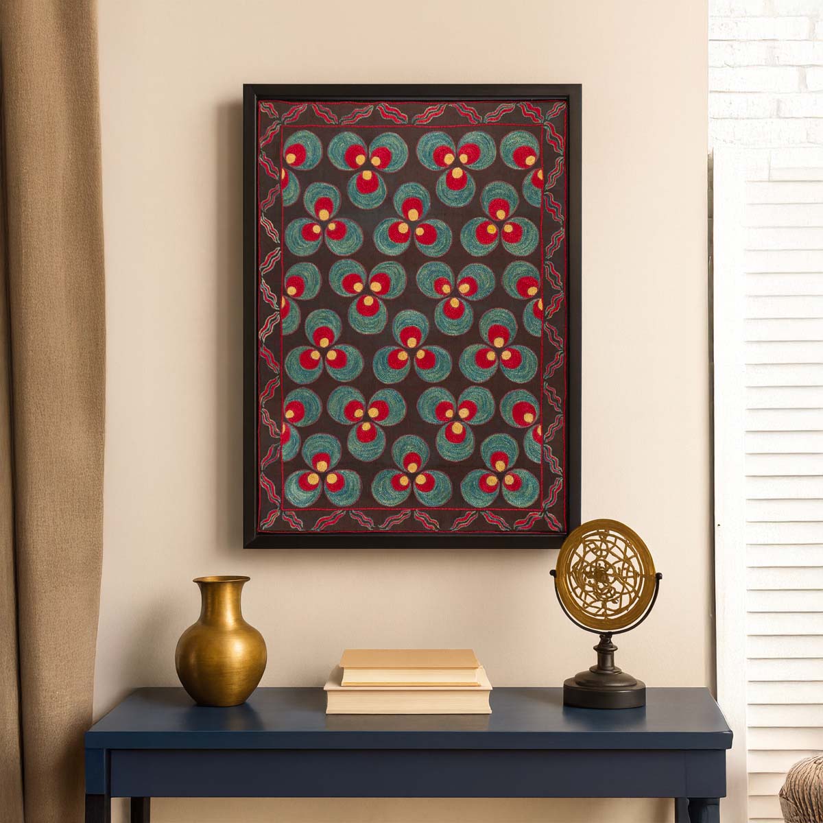In-use interior photo of Mekhann's framed embroidered silk artwork with a Çintamani repeat in teal, red, and ochre on a charcoal ground, set in a matt black frame, hung above a blue console with a brass vase, stacked books, and a decorative brass object.