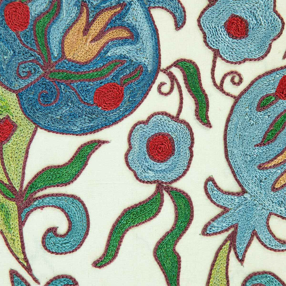 Close up view of Mekhann's hand embroidered cream silk, blue pomegranate artwork. Revealing the embroidered details of the blue pomegranate, surrounded by green leaves and small blue and red flowers.
