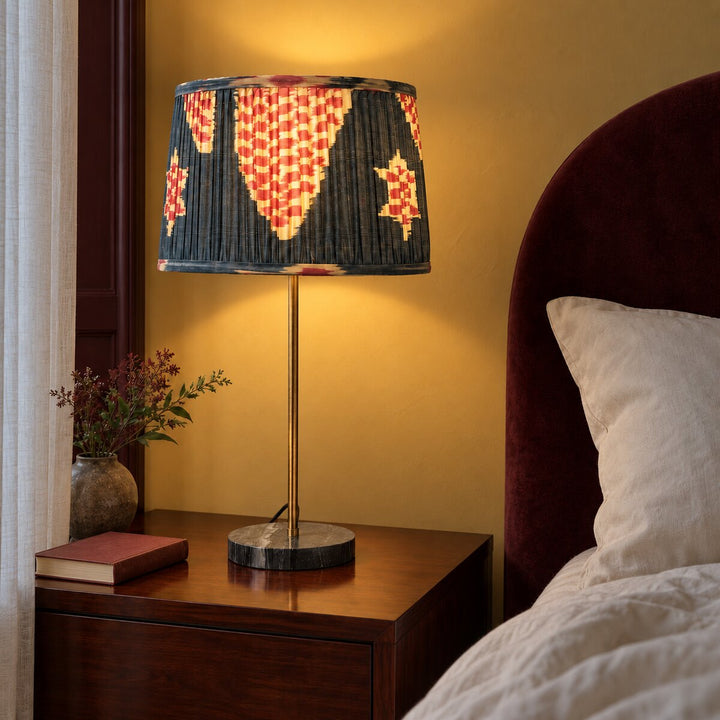 Echo Pewter silk ikat lampshade in Medium, illuminated on a brass bedside lamp beside a burgundy upholstered bed.