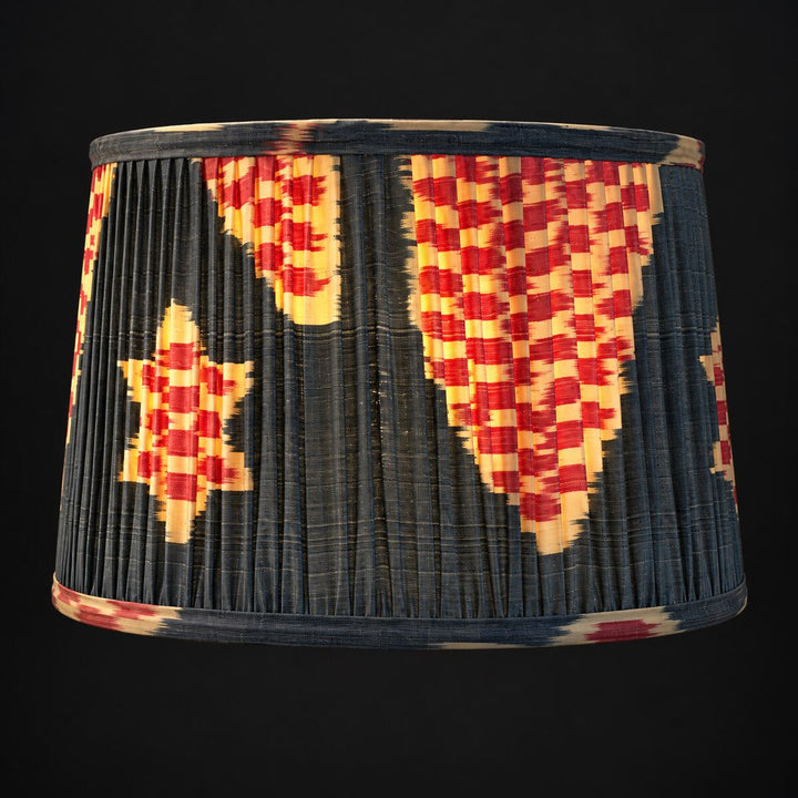Illuminated Echo Pewter silk ikat lampshade in Medium against a dark background, showing the pattern glowing through the pleated silk.