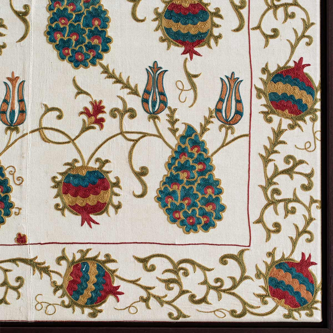 Corner view of Mekhann's cream silk grape vines artwork, showcasing the frame work embroidered onto the actual art in a subtle red tone, within this frame are embroidered pomegranate in red, teal, yellow and red tones.