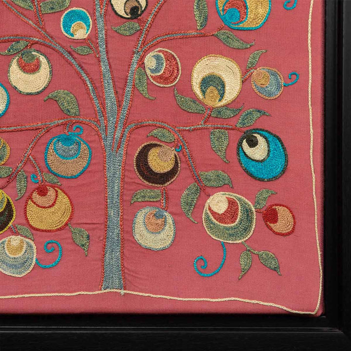 Corner view of Mekhann's pink silk hand embroidered artwork, where we can see the tree of the bottom corner in contrast with the oink silk base and the black frame in view.