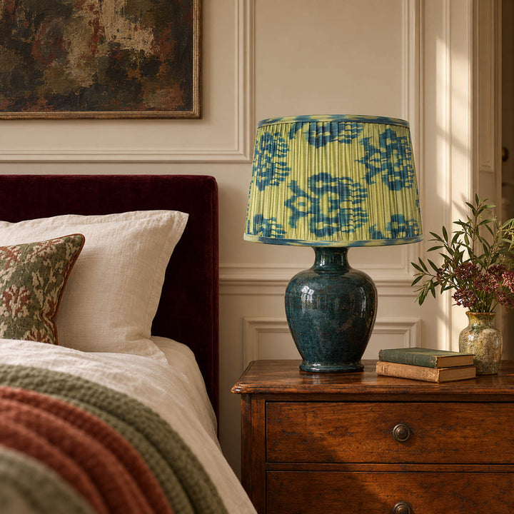 Fleur lampshade styled on a petrol ceramic base beside cream linen and a wine-coloured headboard; illustrative bedroom setting.