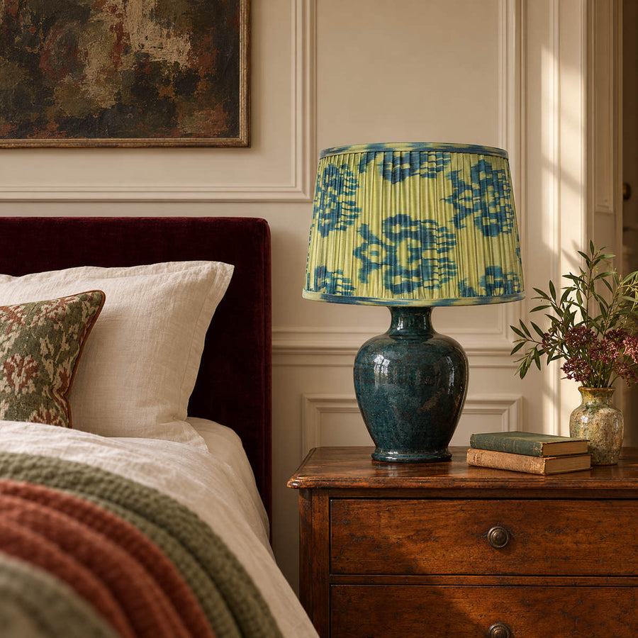 Fleur lampshade styled on a petrol ceramic base beside cream linen and a wine-coloured headboard; illustrative bedroom setting.
