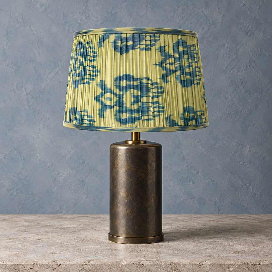 Fleur lampshade styled on a cylindrical bronze-toned base against blue-grey plaster; illustrative full-lamp portrait.
