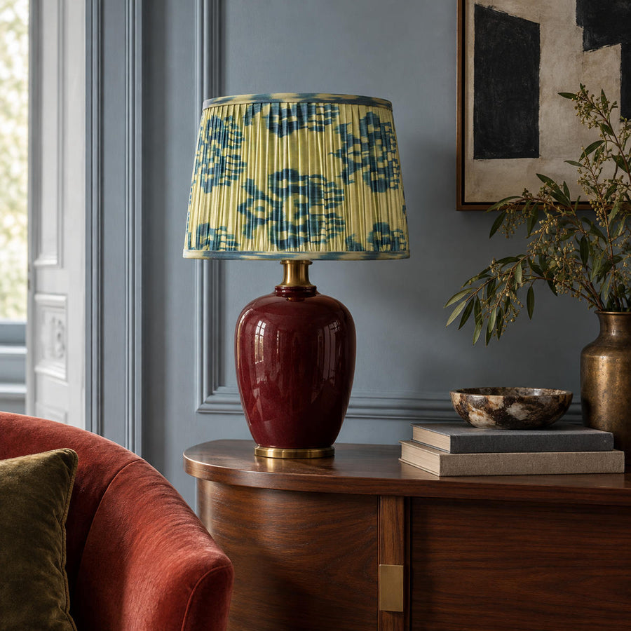 Blue-green Fleur lampshade styled on a burgundy ceramic base against blue-grey panelling; illustrative living-room setting.