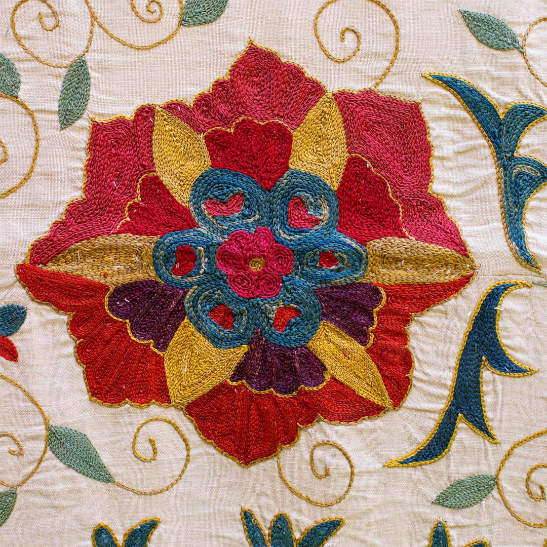 Close up view of the cream bukhara throw, showing one of the many red flower motifs up close. Revealing the detailed intricacies of the hand embroidered flower in red, yellow, blue, purple and pink.
