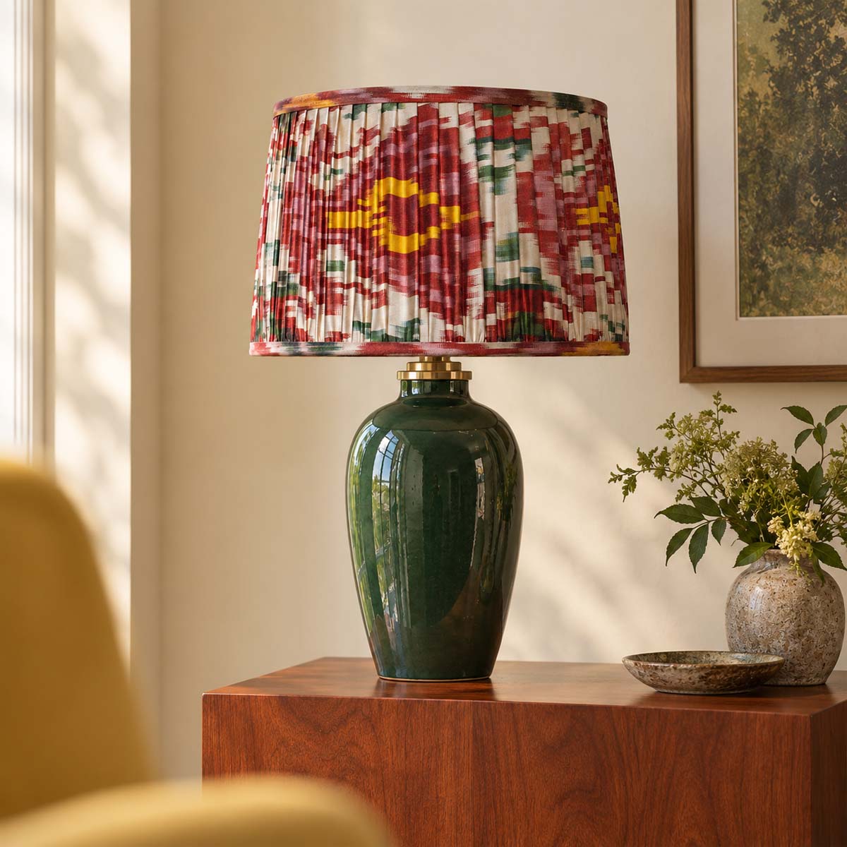 A table lamp with a glossy dark green ceramic base and a pleated drum shade featuring a red, yellow, and green ikat pattern, styled on a wooden side table in a warm, sunlit room.