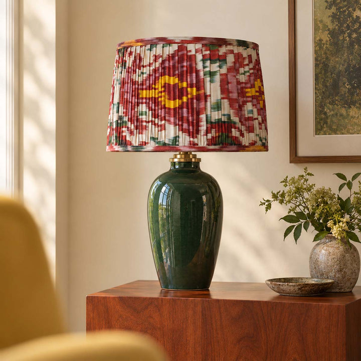 A table lamp with a glossy dark green ceramic base and a pleated drum shade featuring a red, yellow, and green ikat pattern, styled on a wooden side table in a warm, sunlit room.