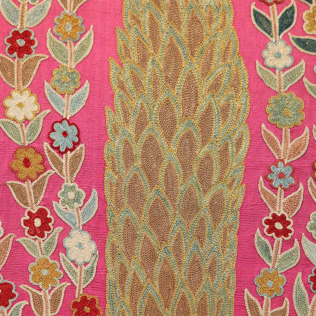 Close up view of Mekhann's pink silk cypress tree artwork, showing the body details of the cypress tree with details of light blue and warm beige silk embroidered. With blue, red, and yellow flowers of each side of the tree, all set on a base of pink silk.