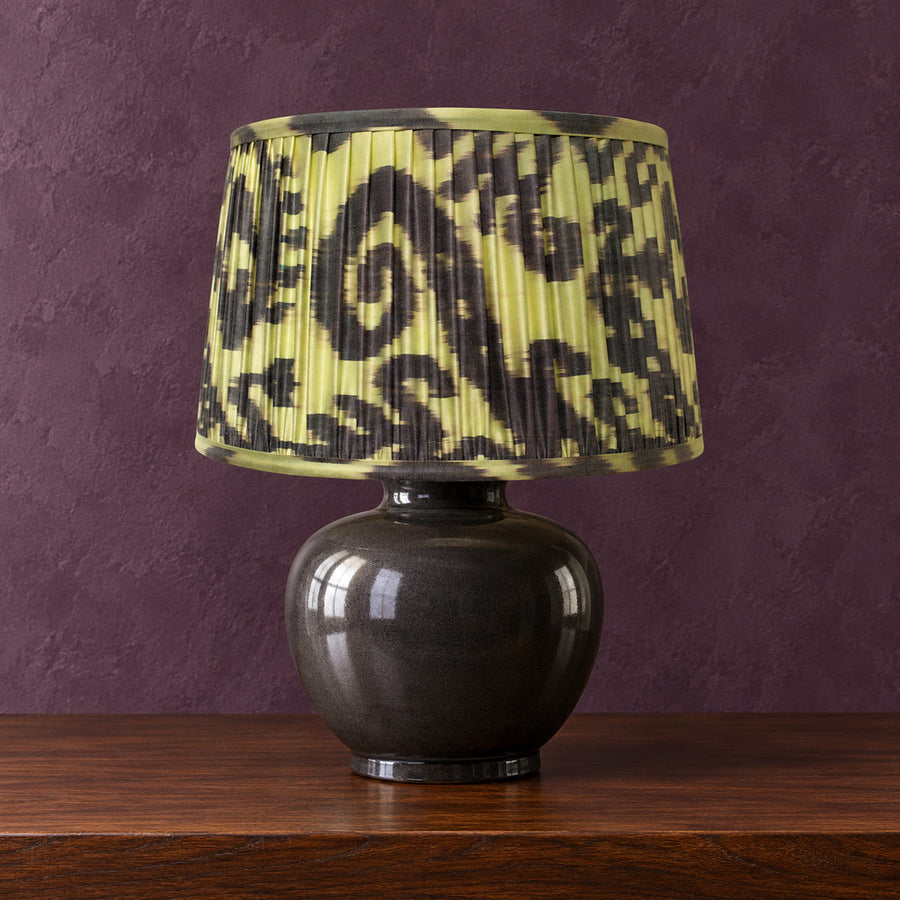 Volute lampshade styled on a dark ceramic base against muted aubergine plaster; illustrative full-lamp portrait.